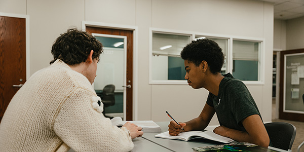 Two students sit at a table in a modern study area, focused on a notebook and textbook while collaborating on a writing project.
